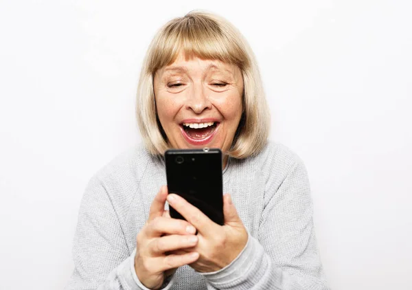 Elég idős nő alkalmi ruhában, modern okostelefonnal és hangosan nevet, miközben fehér háttéren áll. Öreg nő néz vicces videók az interneten. — Stock Fotó