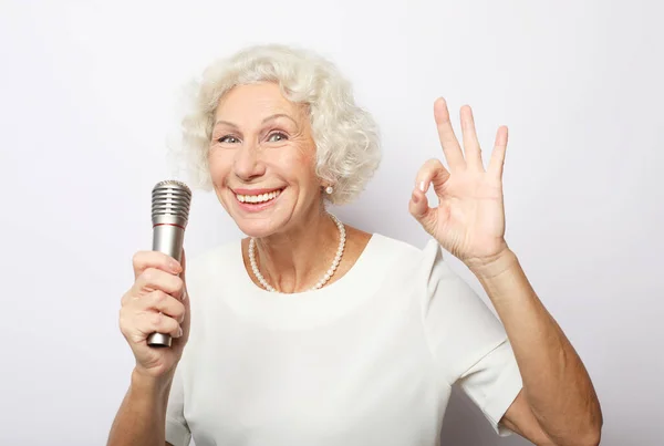 Ευτυχισμένη ηλικιωμένη γυναίκα κρατώντας ένα μικρόφωνο και τραγουδώντας απομονωμένη σε γκρι φόντο — Φωτογραφία Αρχείου