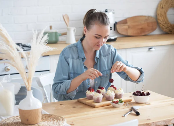 A young woman decorates muffins with white cream in her kitchen. — Stock Photo, Image