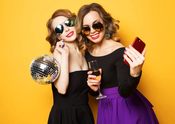 Feier, Party und People-Konzept - zwei modische junge Frauen halten Discokugel in der Hand, trinken Champagner und machen ein Selfie. — Stockfoto