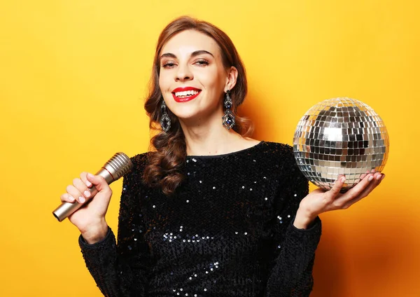 Элегантная брюнетка с длинными вьющимися волосами, одетая в вечернее платье с диско-шаром и микрофоном — стоковое фото