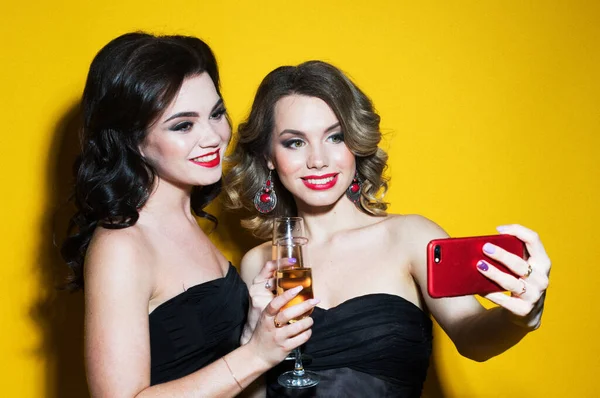 Två chica unga kvinnor i svarta cocktailklänningar dricker champagne och tar en selfie. — Stockfoto