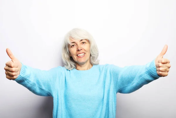 Bom humor feliz sorrindo positivo mulher idosa com cabelos brancos, vestindo suéter azul mostrar polegares para cima. — Fotografia de Stock