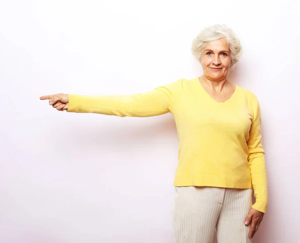 Portrét staré ženy ve žlutém svetru ukazuje, vypadá, ukazuje prsty nahoru kopírovat prostor. — Stock fotografie