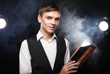 Stüdyoda poz veren profesyonel makyözün elinde gölgeler paleti var. Sigara içmenin arka planına karşı. Genç, modaya uygun bir adam..