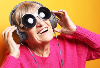 Yaşlılar, tehnoloji ve duygu kavramı: Neşeli yaşlı kadın güneş gözlüğü takıyor sarı arka planda müzik dinliyor