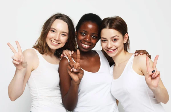Tre unga multiracial kvinnor bär vita skjortor står tillsammans och ler mot kameran, visa v-tecken — Stockfoto