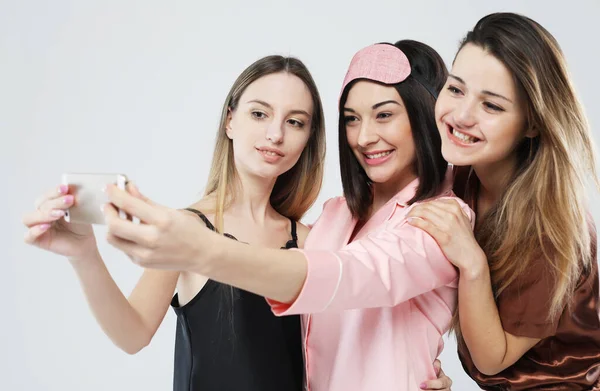 Vänskap, människor och teknik koncept - tre unga kvinnor vänner bär pyjamas med smartphone tar selfie över grå bakgrund — Stockfoto
