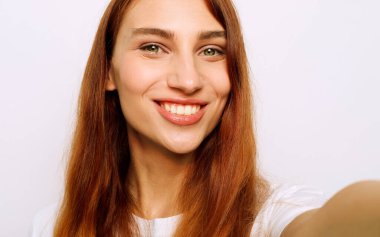 Çekici kızıl kadın cep telefonuyla selfie çekiyor. Güzel genç bir kadın beyaz arka planda poz verirken neşeli bir ifade takınıyor..