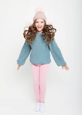 Kıvırcık saçlı, mavi kazaklı ve beyaz arka planda atlayan komik kız çocuğu.