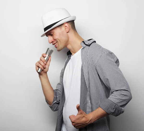 Stile di vita, persone e concetto di svago: un giovane che indossa una camicia grigia e un cappello con in mano un microfono e canta. — Foto Stock