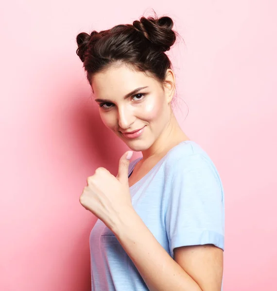 Jonge brunette vrouw in blauw t-shirt glimlacht en heft haar duimen over roze achtergrond. — Stockfoto