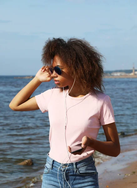 Mladá afro americká žena v slunečních brýlích poslouchat hudbu ve sluchátkách na mobilním telefonu v blízkosti moře. — Stock fotografie