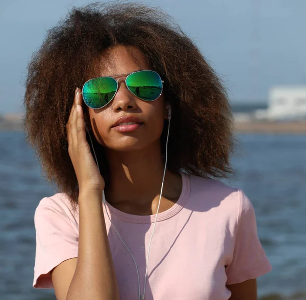 Mladá afro americká žena v slunečních brýlích poslouchat hudbu ve sluchátkách na mobilním telefonu v blízkosti moře. — Stock fotografie