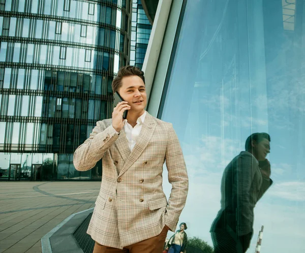 Business, people and success concept: Young handsome businessman talking on his phone outdoors near modern business centre — Stock Photo, Image