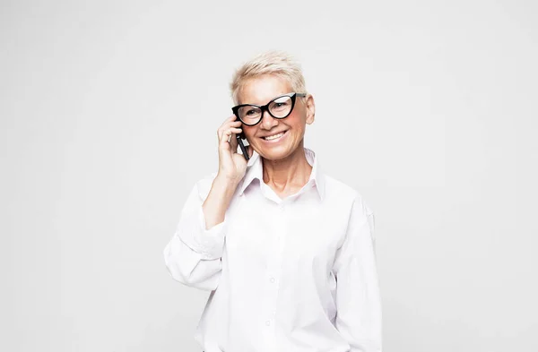 Happy old woman with short hair and glasses using smartphone over grey background. — Stock Photo, Image