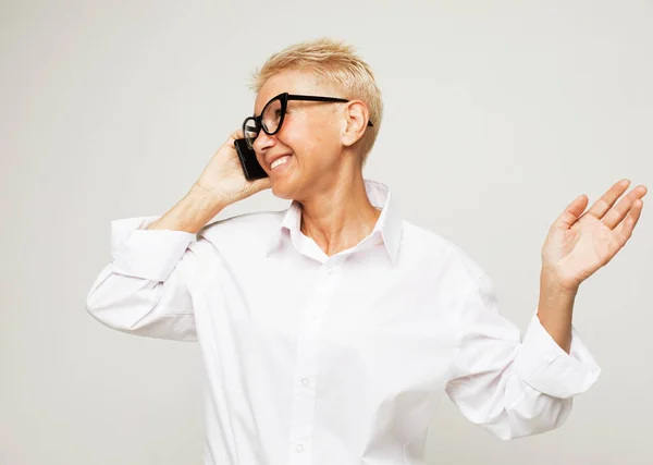 Happy old woman with short hair and glasses using smartphone over grey background. — Stock Photo, Image