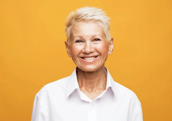 Portrait of happy and charming european senior woman with blond short hair laughing over yellow background — ストック写真