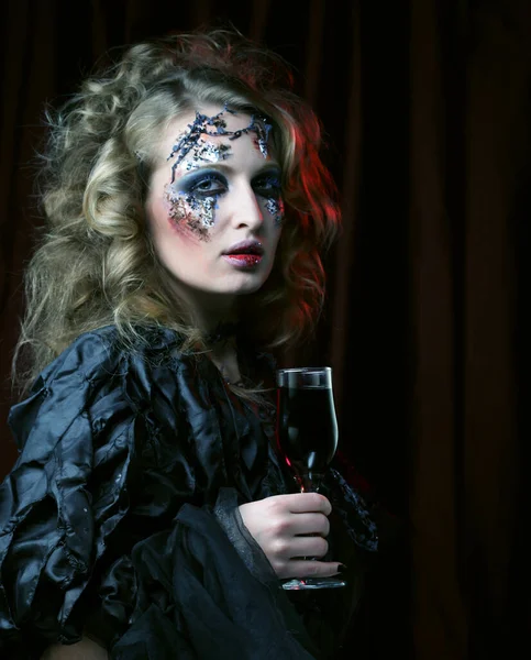 Koncepcja imprezy i Halloween. Piękna kobieta w stroju czarownicy z jasnym makijażem i kręconymi włosami trzyma kieliszek wina. — Zdjęcie stockowe