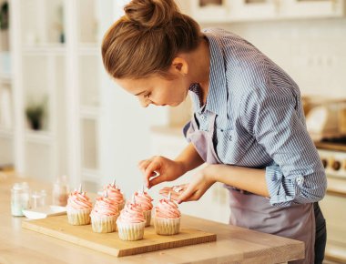 Genç bir kadın muffinleri mutfağında beyaz kremayla süslüyor..