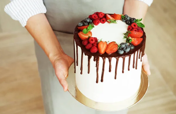 Молодая улыбающаяся брюнетка шеф-повар держит домашний торт украшенный шоколадом и свежими ягодами. — стоковое фото