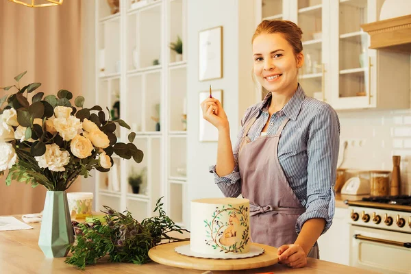 Bella giovane donna decora una torta con la pittura. Un food blogger sta esplorando una nuova ricetta. Home business e nuove opportunità. — Foto Stock