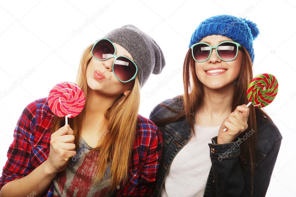 Portrait of two young pretty hipster girls wearing  hats and sunglasses holding candys. Studio portrait of two cheerful best friends having fun and making funny faces.