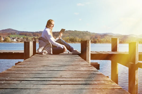 Girl reading from a tablet on the wooden jetty against a lake. S — Stock Photo, Image