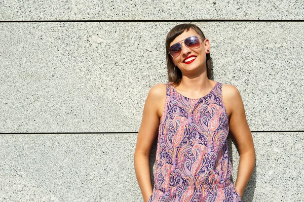 Jeune femme moderne en lunettes de soleil près du mur en ville souriant à — Photo