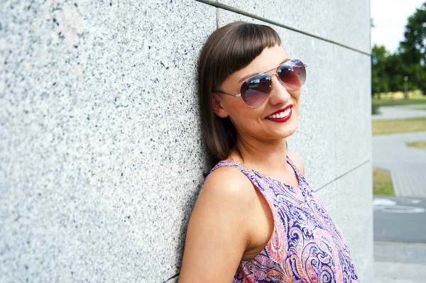 Jeune femme moderne en lunettes de soleil près du mur en ville souriant à — Photo