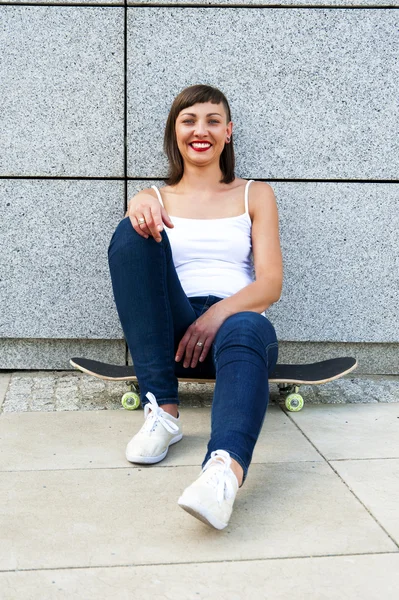 Jovem menina moderna sentar-se no skate pela parede na cidade . — Fotografia de Stock