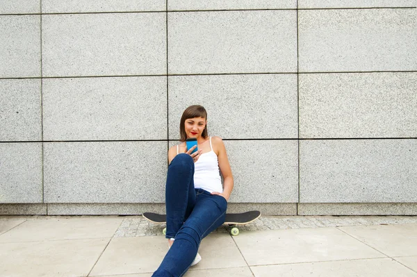 Jeune fille assise sur skateboard dans la ville avec téléphone par la wa — Photo