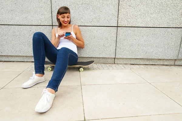 Niña sentada en el monopatín en la ciudad con el teléfono por la wa — Foto de Stock