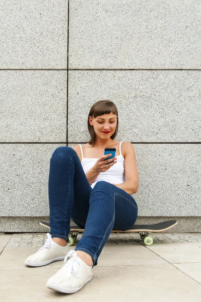 Niña sentada en el monopatín en la ciudad con el teléfono por la wa — Foto de Stock