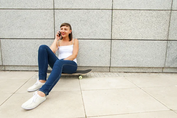 Niña sentada en monopatín en la ciudad hablando por teléfono nea —  Fotos de Stock