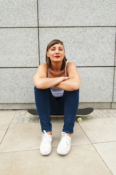 Молодая современная девушка сидит на скейтборде у стены в городе . — стоковое фото