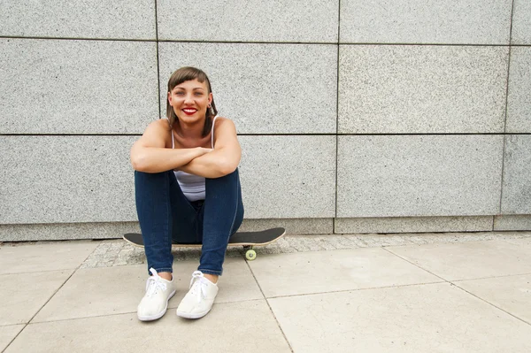 Giovane ragazza moderna sedersi sullo skateboard vicino al muro in città . — Foto Stock