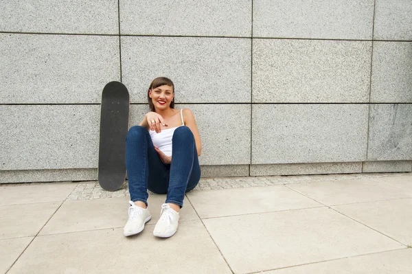 Junges Mädchen mit Skateboard an der Mauer in der Stadt, lächelnd. — Stockfoto