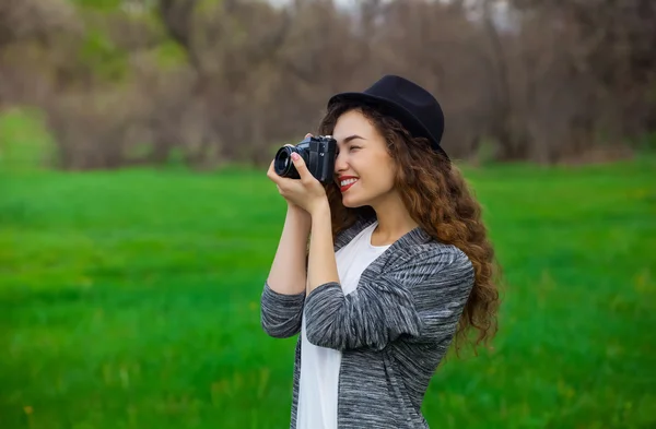 Menina jovem, bonita, sorridente em um chapéu e com longas, cabelo encaracolado fotos da natureza no filme do parque, câmera SLR . — Fotografia de Stock