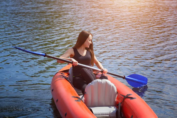 Den smilende pige sportskvinden med langt, mørkt hår i sort, sportstøj rækker med en åre på søen i en rød, oppustelig kano i en varm, sommer, solrig dag. Beskæftigelse ved sport roning på en kajak . - Stock-foto