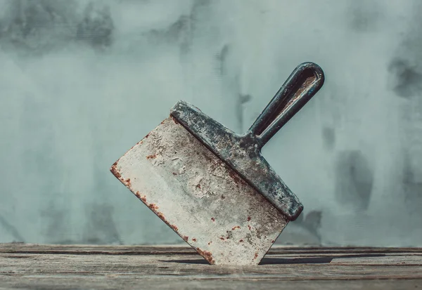 In the studio,on the background of plastered walls,stuck the blade down into the wood,cracked,worktop vintage,used,rusty,smeared in a mortar,darkened,steel,metallic,dirty,work tool spatula. — Stock Photo, Image