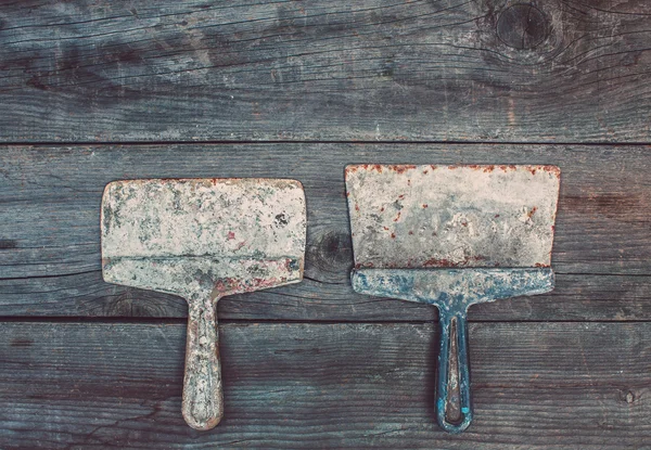 On the old, wooden, cracked, the working surface in the studio are two vintage, used, rusty, darkened, mettalic, smeared with mortar instruments of different sizes - spatulas. — Stock Photo, Image