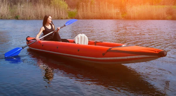 Den smilende pige sportskvinden med langt, mørkt hår i sort, sportstøj rækker med en åre på søen i en rød, oppustelig kano i en varm, sommer, solrig dag. Beskæftigelse ved sport roning på en kajak . - Stock-foto