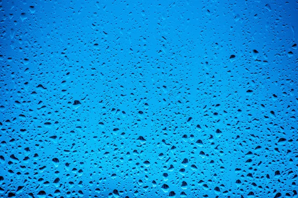 Water drops flow down on the glass in the rain, on a blue background — Stock Photo, Image