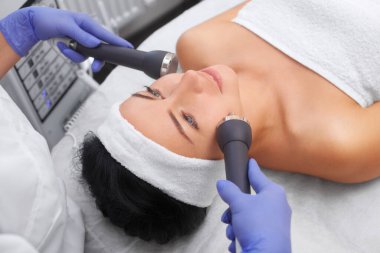 The cosmetologist makes the procedure an ultrasonic cleaning of the facial skin of a beautiful, young woman in a beauty salon.Cosmetology and professional skin care. clipart