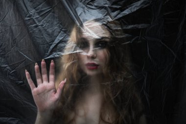 Genç, güzel, üzgün kadın COVID-19 'a karşı korunmak için plastik bir çarşafın arkasında mahsur kaldı. Tam kitap kapağına göre. 