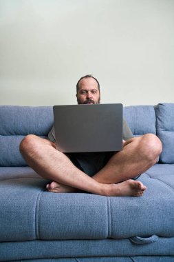 Bir adam kanepede bağdaş kurup dizüstü bilgisayarına odaklanmış oturuyor. Basit duvarları olan bir odada..