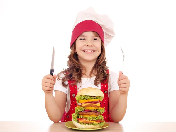Glückliches kleines Mädchen kocht mit großem Hamburger auf dem Tisch — Stockfoto