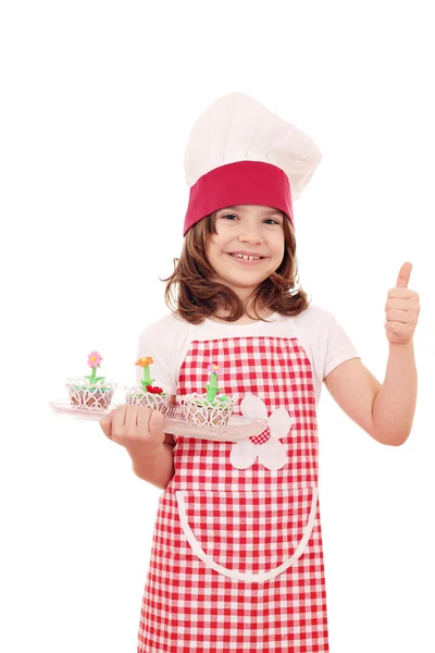 Happy little girl cook with cupcakes and thumb up — Stock Photo, Image