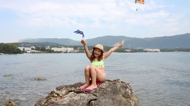 Menina feliz sentada em uma rocha junto ao mar e acenando bandeira grega — Vídeo de Stock